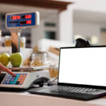 Digital laptop displaying white screen is positioned on checkout counter of eco friendly store. While storekeeper weighs apples on scale, the wireless computer shows blank copyspace mockup template.